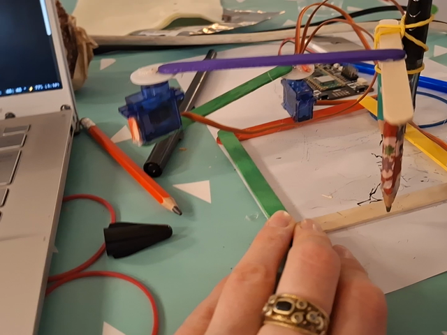 Robot made of small servo motors and lollipop sticks with a pen attached drawing chaotic lines on a sheet of paper