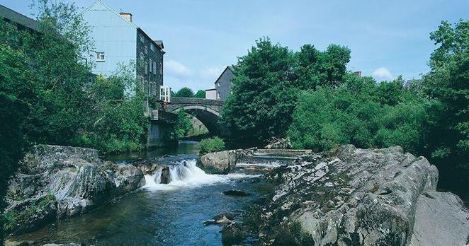 A photograph of the river Wye running through the town of Rhayader. It tumbles over rocks and is both spectacular and peaceful. The sky is blue with very few clouds.