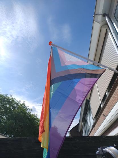 A Progress Pride flag suspended from a short pole at the front of a house, with a blue sky in the background