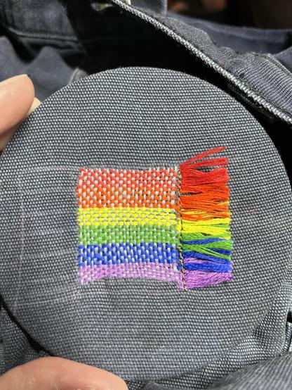The mended pants have a square rainbow flag with a little rainbow fringe to the right hand side. The horizontal stripes are colored, and the cross-weave is a dark grey that blends in to several of the colors.