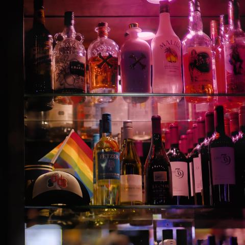 stylized photo taken inside a bar. Many glass bottles of liquor are backlit on glass shelves. In the bottom left a pride flag is discreetly displayed next to a logo-ed baseball hat