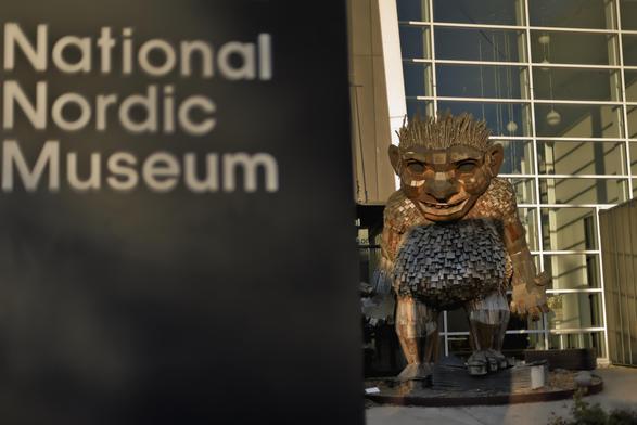 the frame is divided roughly in half by a large black free-standing sign in the foreground that simply reads "National Nordic Museum" in plain lettering. The right half shoes a glass and metal building with a large atrium behind a wooden sculpture of a troll approx 3 meters tall.