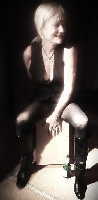 blond white woman in metal chick outfit (me), sitting astride a cajon, beating along unheard music. She smiles to an unseen band member, Colors are tuned down, shadows rather dark. An empty bottle of beer shares the limelight