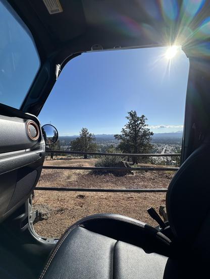 View from the driver’s seat of a big hole where the passenger door would be, sun with lens flare peeking in at the top. Juniper trees and scrub in the foreground, city streets and mountains beyond in the background. Soft blue sky with very few clouds.