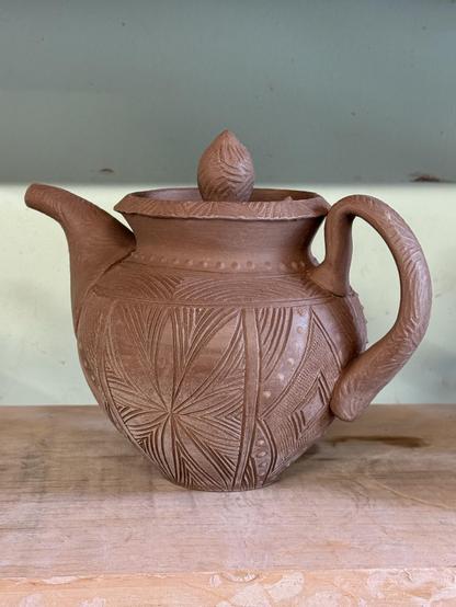 A handcrafted clay teapot with intricate geometric patterns and a decorative lid sits on a wooden shelf. The teapot features a curved handle and a spout, both with subtle surface texture. The teapot is at the leather-hard stage—a rich brown color. It has not been fired.