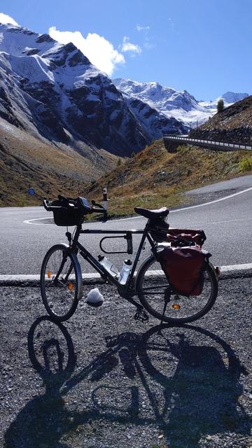 Fahrrad mit Lenkertasche und roten Packtaschen am Straßenrand abgestellt.
im Hintergrund ein grandioses Bergpanorama.