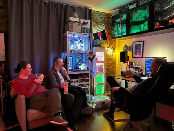 Liz, Terence and myself in a dark room with much retrocomputing: A makeshift rack with a 486 "server", some large LED-equipped floppy disks on the wall, Day Of The Tentacle poster on another wall, the screen of a computer running DOS, and several cupboards with colourful lights and various retrogear.