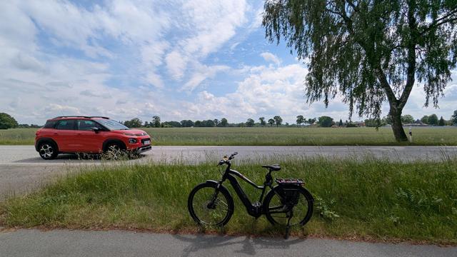 Ein schwarzes Haibike Trekking Fahrrad steht auf einem Fahrradweg in Fahrtrichtung nach links. 
Auf der dahinterliegenden Bundesstraße fährt ein rotes Auto in Fahrtrichtung nach rechts. 
Eine Birke steht am Straßenrand.
Der Himmel ist leicht bewölkt.