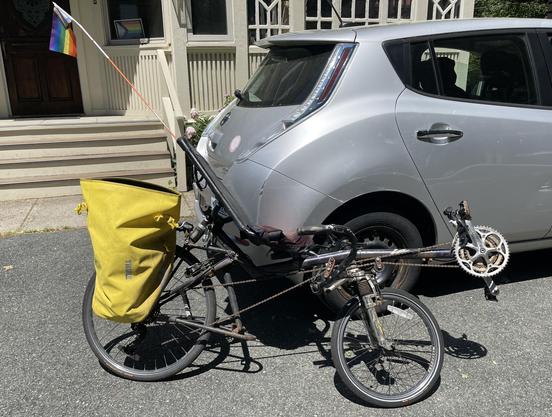 A black recumbent bicycle with a short wheelbase (gears/pedals out in front) and under seat steering leaning against a silver Nissan Leaf in front of a white house. The bicycle has a yellow pannier on the back and a progress pride flag sticking out of the top of the seat. If you squint, you can barely make out a progress pride flag in a window of the house.