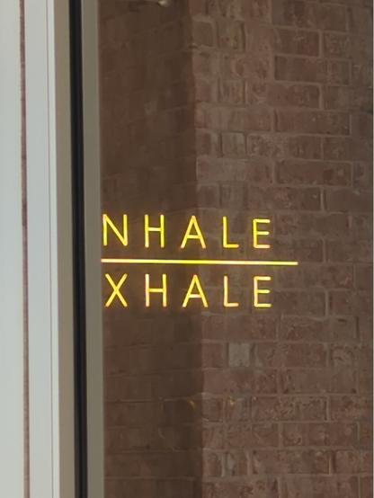Artistic rendition of a yellow faux-neon INHALE / EXHALE sign inside a yoga studio. The I and the E are obscured by the aluminum door frame, and the interior of the studio is obscured by the reflection of the brick column outside, so it looks as if the sign is mounted on brick, and half of the letters are hanging out in space.