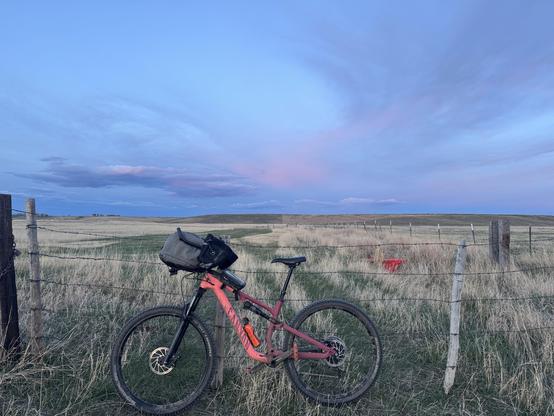 A big pink mountainbike leaning against a farm gate framed by high evening clouds. A bicycle is the happiest way.