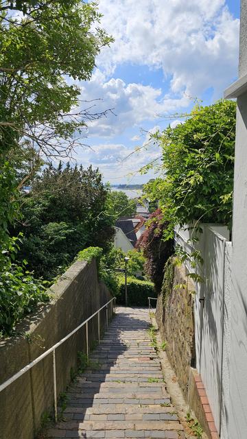 Schmale Treppe mit Pflastersteinen zwischen einer Mauer links und einer Steinmauer mit weißem Zaun rechts. Beidseitig dichter Bewuchs mit Bäumen, Sträuchern und Kletterpflanzen. Im Hintergrund sind Dächer von Häusern sowie ein Gewässer sichtbar. Darüber blauer Himmel mit großen Wolken.