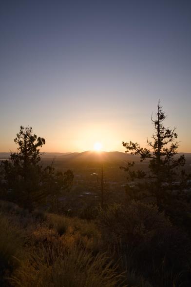 Sun peeking above mountains in the distance, town in the middle ground, juniper and scrub grass faintly illuminated in the foreground.