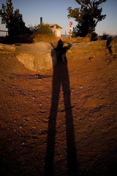 Elongated shadow of me holding camera to face, against small pebbles of the trail, some bigger rocks, and the top-of-butte bathroom and stop sign and juniper trees.