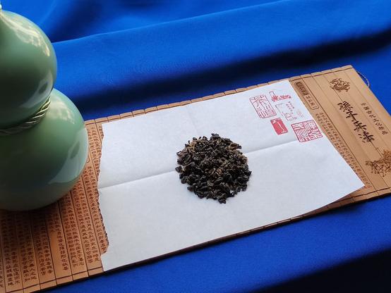 Today's tableau features a blue backdrop cloth, an unfurled bamboo scroll of 孙子兵法 (Sūnzǐ Bīngfǎ, "Sun-Tzu's The Art of War"), and a celadon-coloured calabash-shaped bottle.  A piece of torn paper folded two ways to help center the tea piled in its middle covers much of the scroll.  A collection of red seals is stamped on the top right for decoration.

This first view of the tea shows the tightly curled dark leaves, with visible trademark orange-tipped buds and fine hairs.