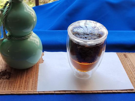 Now the bulk of the work is being done by the water.  A double-walled, lidded glass with a glass basket holding the leaves is filled to the top with the umber liquor—the reason why the Chinese call this tea "red" (in English we describe the leaf as "black").  The leaves have fully unfurled and fill the basket, with plenty of space between them for water to circulate.  Under the basket, at the bottom of the glass, the pure liquor is visible against the white paper background which beautifully contrasts the colour of the tea.

To the left of the tea is again the celadon-coloured, calabash-shaped bottle.
