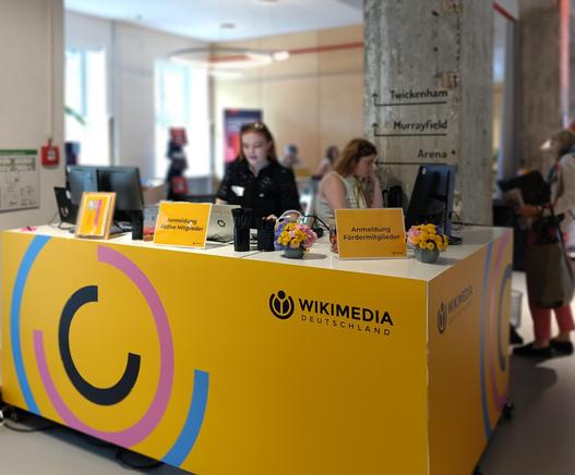 Einladender Empfangstresen zur Anmeldung, dekoriert mit Wikimedia-Logos, Leitsystem und Namensschildern einer Mitgliederversammlung, Personen im Hintergrund sind unscharf.