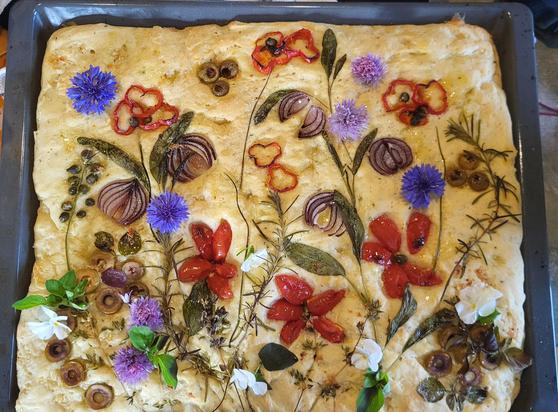 Ein rechteckiges Fladenbrot in einer dunkelgrauen Backform ist mit verschiedenen Zutaten wie ein Blumenarrangement angeordnet. Auf dem hellen Teig sind halbierten Tomaten, Oliven, Zwiebelringe und grüne Kräuterzweige so platziert, dass sie Blüten und Stiele imitieren. Kleine violette und blaue Blüten sind über das Brot verteilt.