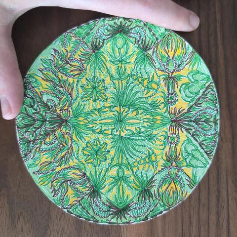 Circle-shaped paper cut-out with intricate patterns drawn on it (mostly leaves and pearls). Colours are green, brown, and yellow. Background is a walnut wood table.