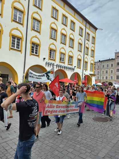 Demo-Zug durch die gepflasterte Fußgängerzone einer Altstadt: Die Gruppe, die gerade vorüberzieht, hat eine rote Linke-Fahne dabei und trägt ein Transparent in Regenbogenfarben, auf dem steht: „Alle Geschlechter gegen den Faschismus“. Auf einem anderen Transparent steht: „The first Pride was a Riot!“. Die folgende Gruppe trägt ein großes Regenbogentransparent vor sich her. Auf dem historischen Gebäude dahinter steht „Café Bergmeister“.