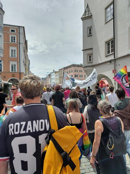 Pride-Demozug durch eine Altstadt, aus dem Zug heraus fotografiert. Rechts und links sind die renovierten Fassaden von mehrstöckigen historischen Gebäuden zu sehen, dazwischen geht ein schier endloser Zug von Menschen, die Transparente und Fahnen tragen. Die Leute sind von hinten zu sehen. Eine Fahne kombiniert den Regenbogen mit den schwarz-roten Antifa-Fahnen. Ein Mensch ganz vorne trägt ein dunkles T-Shirt mit der Rückenaufschrift „Rozanov 81“.