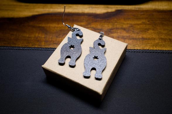 Two 3D-printed earrings sitting on a small box. Each is an identical silhouette of a cat from behind, with the tail curling up to meet the earring hook and clasp. In the middle of each, and rather prominent, is a butthole asterisk.