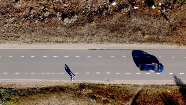 overhead view of asphalt road with white dashed stripes on each edge, a car is driven along the center lane as a person rides a bike on the edge lane