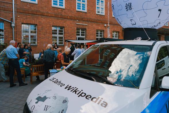 Vordergrund: Bus mit der Aufschrift "25 Jahre Wikipedia", auf dem Dach des Autos ist ein großer Ballon. Im Hintergrund: Menschen stehen uns sitzen an Tischen.