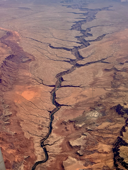 aerial view of a desert canyon landscape,  photographed from an airplane window. A narrow river winding through a vast expanse of red and tan sandstone, carving deep, dramatic canyons into the plateau.