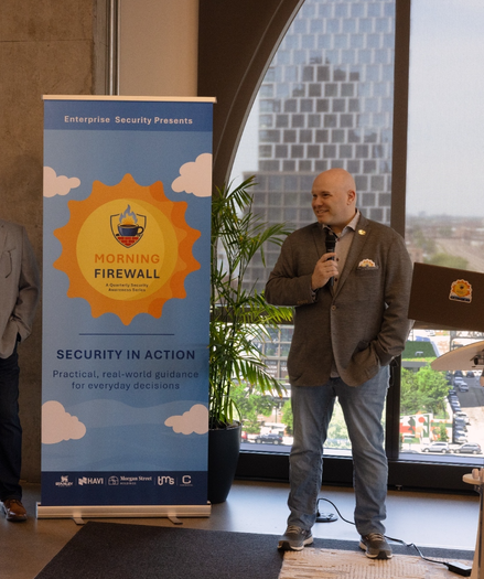 Photo of a man in a gray jacket with a shiny lapel pin standing next to signage for an event. The Event Signage reads Morning Firewall Security In Action. Practical, real-world guidance for everyday decisions.
