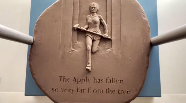 Image of round, wooden or plaster plate, inspired by an image of the famous '1984' commercial from apple, whereby an athletic woman in racing outfit with shorts is running towards the eye of the beholder, holding an heavy slashhammer. The figure, holding the hammer is precisely cut our (wood) or baked, modeled on top of the round plate.  The text under the figure reads 'The Apple has fallen so very far from the tree'.