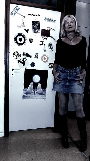 photo showing a caucasian woman (me) posing at the side of a door full of stickers. She is wearing a short jeans skirt, a black shirt baring her neck and part of her collarbones. Amongst other things the colours are manipulated to nearly drown in darkness, to make the picture gloomy, besides the bright door. Nonetheless the details ate still nice to see, some structures of the cork floor, the structure of her hair, her deeply tanned hands and shoulders,