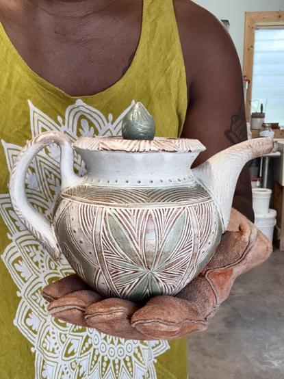 A closeup of me holding a teapot with my hand protected by a large orange kiln glove. I’m wearing a light olive green dress with a white mandala design and you can see a bit of my studio in the background.  

The teapot is white with light blue accents and lots of carved geometric line patterning and stamped detail.
