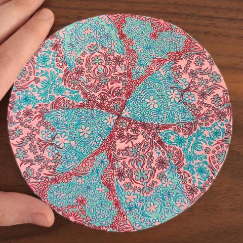 Circle-shaped paper cut-out with intricate patterns (various floral and leafy shapes) drawn on it using fineliners (colours are wine red, coral pink, turquoise, jade). Background is a walnut wood table. You can also see my thumb and index finger in the left corners; the circle is roughly the size of my hand.