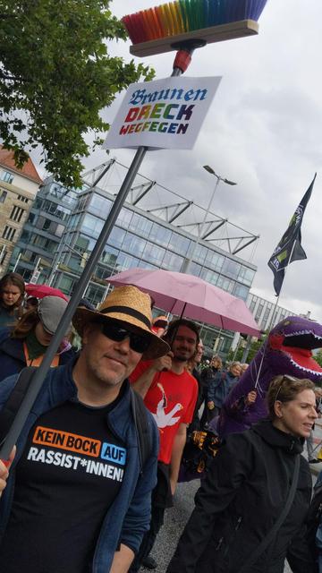 Bild von der Prüf Demo Hannover. Mann mit Schild "braunen Dreck wegfegen" an Regenbogenbesen.