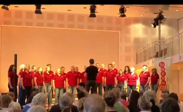 Standbild aus einem Videoschnipsel vom Auftritt des Chors "Unerhört" aus Dortmund unter der Leitung von Christian Koops. Auf einer Bühne vor holzvertäfelter Wand steht ein gemischter Chor von 22 Personen, annähernd paritätisch Frauen und Männer. Der Chor trägt weinrote Polohemden und Jeans, der Chorleiter ist ganz schwarz angezogen und steht mit dem Rücken zum Publikum und zur Kamera. Die Sitzreihen in Vordergrund sind fast vollständig besetzt.