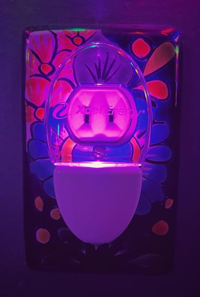 A small, low power, oblong-shaped, LED night light plugged into a wall outlet covered by a decorative ceramic tile. The night light is glowing purple and has the name of the utility company printed on the clear plastic light covering.