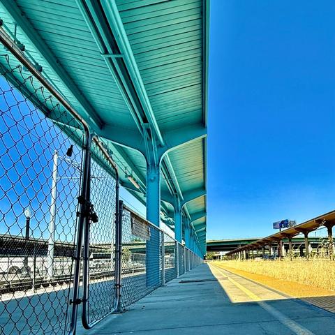 Defunct train platform on a sunny day. The tracks are overgrown And the area is blocked off with a chain link fence.