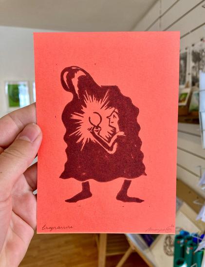 A photo of my hand holding up a linocut on pink fluorescent paper, printed in a dark burgundy ink, sized A6. It shows a screaming ghost, bended over with inside of him a little figurine with a light, shining brightly.