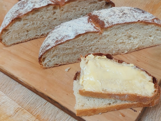 Two stacked slices of a milk loaf, one of them buttered. Behind are the two halves of the loaf, with a floured thin brown crust.