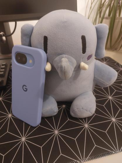 A blue mastodon plushie holding a pixel 10a in lavender color. Taken with a Samsung A52s.