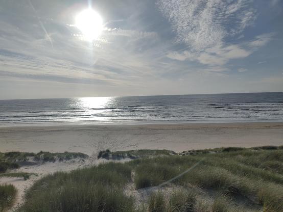 The sun over the north sea. It's fairly low, but not yet setting. Below the dune grass gives way to a sandy beach. Small waves are crashing onto the beach.