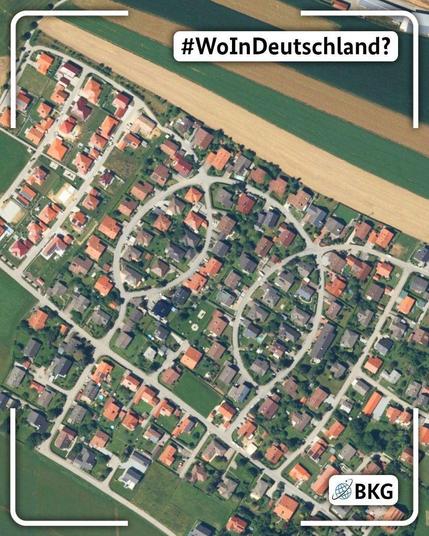 Luftbild eines Wohngebiets mit Einfamilienhäusern, Straßen und angrenzenden Feldern. Die Straßenführung bildet auffällige kreisförmige Muster innerhalb der Siedlung. Über dem Bild steht die Frage „#WoInDeutschland?“, unten rechts ist das BKG-Logo eingeblendet.