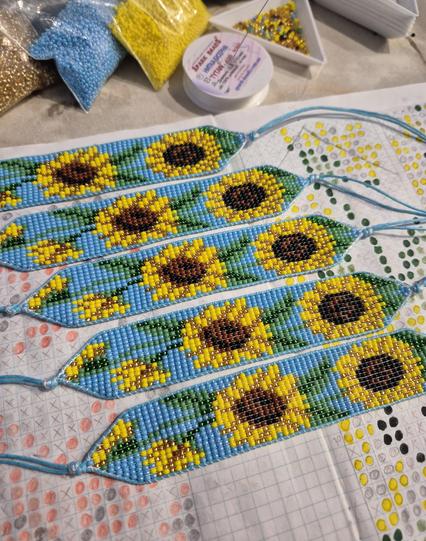 ​Five handmade beaded bracelets with a pattern of vibrant yellow and brown sunflowers on a bright blue background. The bracelets are arranged neatly on top of a hand-drawn beading pattern sheet. In the background, bags of blue, yellow, and gold seed beads are visible alongside a spool of white thread.
