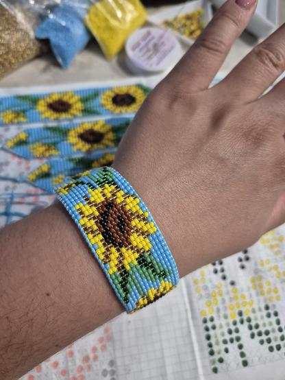 A close-up shot of a person's wrist wearing a wide, handmade loom-beaded bracelet. The bracelet features a detailed pattern of bright yellow and brown sunflowers with green leaves woven onto a sky-blue background. In the blurred background, other similar beaded bracelets, a hand-drawn pattern sheet, and bags of loose seed beads are visible.