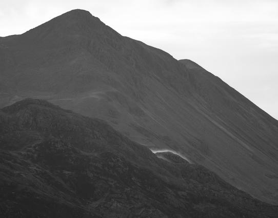 Monochrome photo. A small,, isolated wave of mist, lit by the sun, flows over a rocky bulge on a huge mountainside.