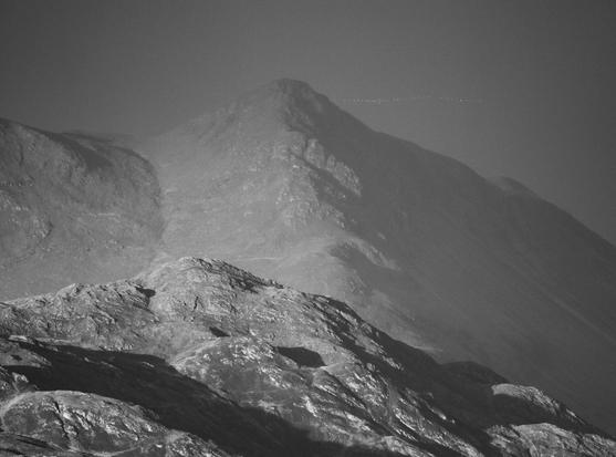 Monochrome photo. A mountain is lit by the evening sun. Just to the right of the peak a distant flock of geese, one behind the other in single file, flies into the sun.