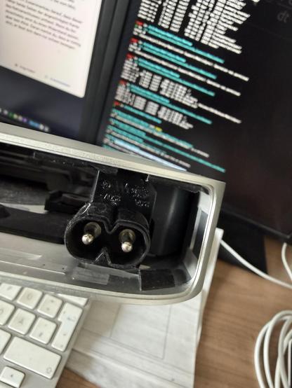 Es zeigt einen elektrischen Anschluss mit zwei Metallstiften, der an einem Metallgehäuse befestigt ist. Im Hintergrund zeigt ein Computermonitor Codezeilen oder Daten an, und im Vordergrund ist eine Tastatur zu sehen.