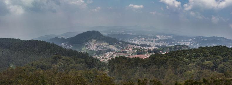 View of a town spread across a few hills from an elevated hiking trail in the distance. Forest canopy in the foreground, cloudy sky against distant hills in the background