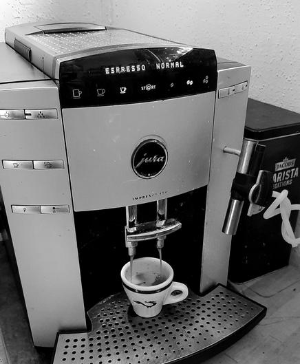 Black and white photo of my coffee maker, in the process of producing an espresso in medium strength. 

A boxy aluminium housing with a company logo, an array of buttons, an outdated digital display with letters by lighted dots, the froth maker with its white tube knotted at the side, and a tin box of beans to the side. A small espresso cup on a shiny metal grad is being filled.